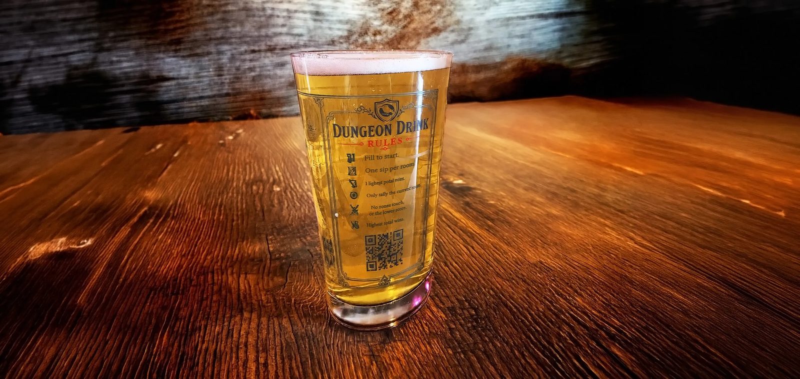A filled Dungeon Drink pint glass on a wooden bar top, the etched rules panel and its QR code facing the camera